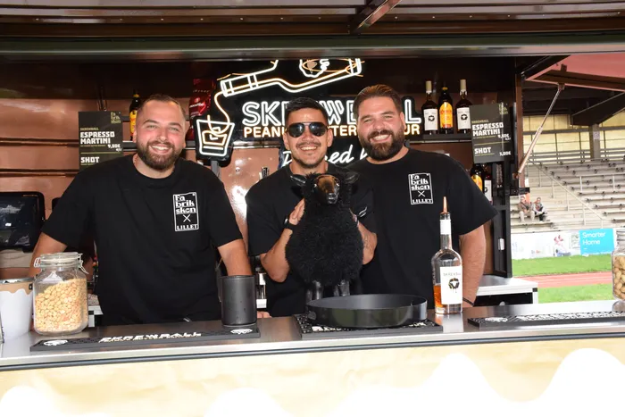 Drei lächelnde Männer hinter dem Tresen der Bar „Skrevell“, einer von ihnen hält ein kleines schwarzes Schaf.