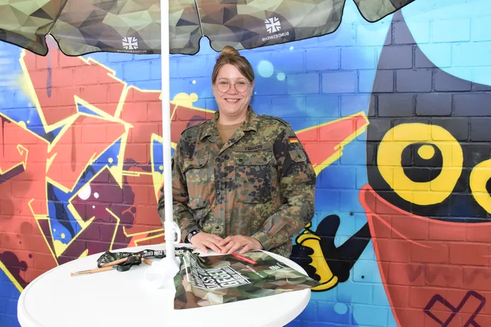 Eine lächelnde Frau in Bundeswehr-Uniform mit dem Namensschild „Verhoff“ steht vor einer bunten Graffiti-Wand.