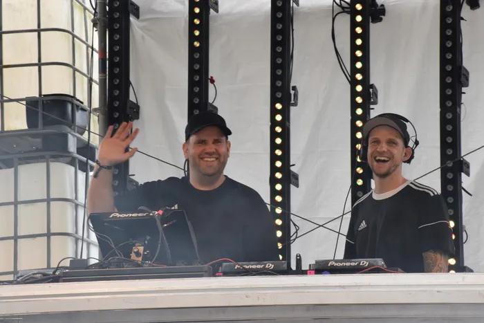 Zwei lächelnde Männer hinter einem DJ-Pult mit Pioneer-Equipment, einer von ihnen winkt.