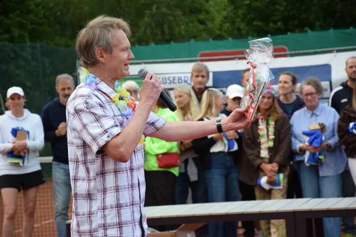 Ein Mann mit einer Blumenkette hält ein Mikrofon und präsentiert ein eingepacktes Geschenk bei der Jubiläumsfeier der MTC/TC Halver Open.