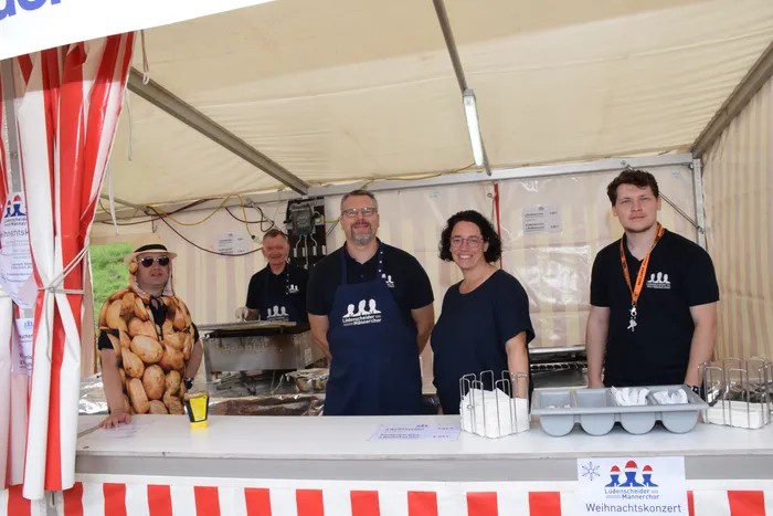 Vier Männer des Lüdenscheider Männerchors stehen hinter der Theke eines Imbissstandes unter einem Zelt; einer trägt ein Hemd mit Brötchen-Motiv.