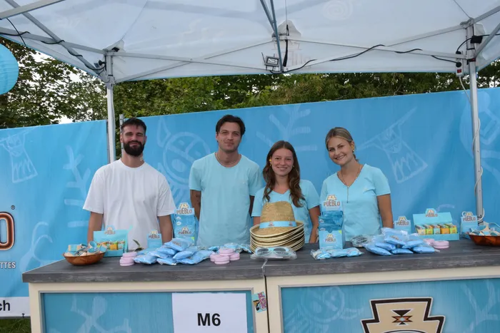 Vier lächelnde Personen stehen hinter einem blauen Stand mit Pueblo-Produkten und Hüten unter einem weißen Zelt.