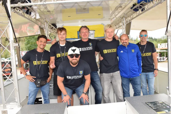 Sieben Männer, die meisten in schwarzen „BEN SPORT“-T-Shirts, posieren lächelnd unter einer weißen Überdachung.
