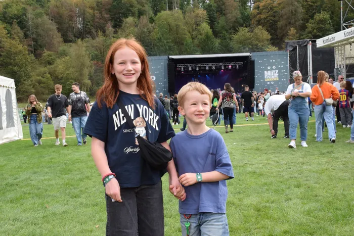 Ein rothaariges Mädchen und ein Junge halten Händchen auf einer Wiese vor einer Festivalbühne und einer Menschenmenge.