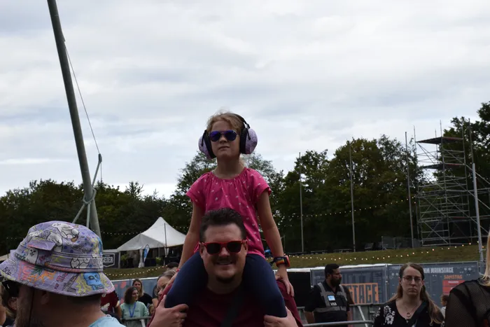 Ein Mädchen mit Sonnenbrille und Kopfhörern sitzt auf den Schultern eines Mannes bei einer Open-Air-Veranstaltung.