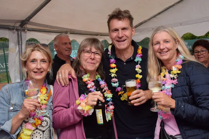 Eine Gruppe lächelnder Menschen mit bunten Blumenketten hält Gläser mit Bier und Wein unter einem Festzelt hoch.