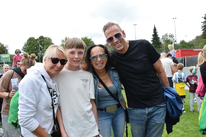 Vier lächelnde Personen posieren für ein Gruppenfoto auf einer Wiese bei bewölktem Himmel.