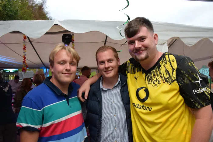 Drei lächelnde Männer posieren unter einem weißen Zelt, einer trägt ein gelbes BVB-Trikot.
