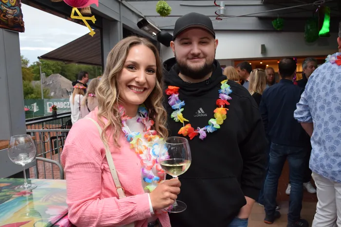 Eine lächelnde Frau in einem rosa Oberteil und ein Mann in einem schwarzen Kapuzenpullover tragen bunte Blumenketten. Die Frau hält ein Glas Weißwein.