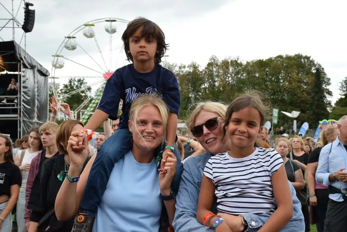 Eine Frau trägt ein Kind auf den Schultern, daneben lächeln eine weitere Frau und ein Mädchen vor einem Riesenrad bei einem Festival.