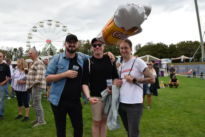 Drei lächelnde Männer stehen bei einem Open-Air-Event vor einem Riesenrad, über ihnen schwebt ein großer Ballon in Form eines Gebäckstücks.