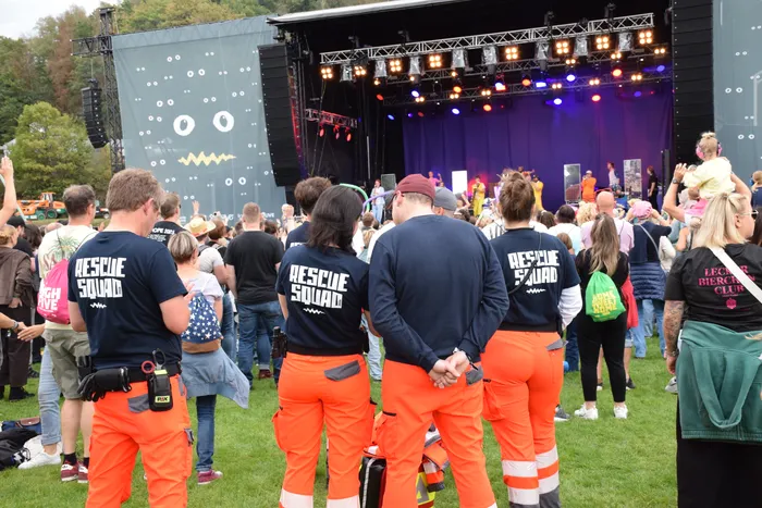 Mitglieder der Rescue Squad in schwarzen T-Shirts und orangefarbenen Hosen stehen in einer Menschenmenge vor einer beleuchteten Bühne bei einem Open-Air-Event.