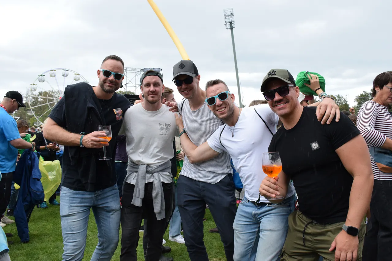 Eine Gruppe von fünf Männern posiert lächelnd bei einem Outdoor-Event, im Hintergrund ist ein Riesenrad zu sehen.
