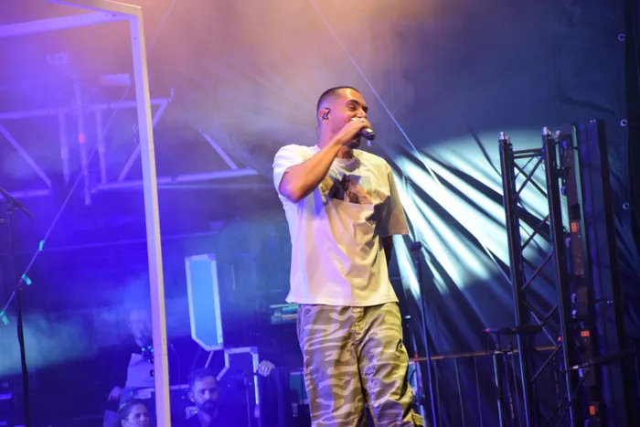 ClockClock singt in ein Mikrofon, bekleidet mit einem weißen T-Shirt und Camouflage-Hosen, unter blauem Bühnenlicht.