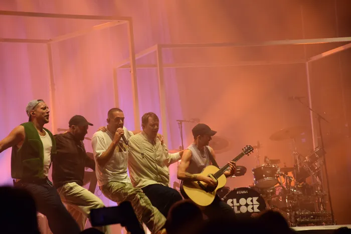 Die Band ClockClock tritt auf einer Bühne unter orangefarbenem Licht auf; ein Sänger hält ein Mikrofon und ein Musiker spielt Akustikgitarre.