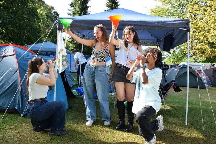 Vier junge Menschen lachen und spielen mit bunten Trichtern auf einem Campingplatz zwischen Zelten.