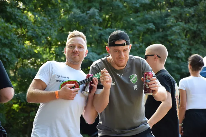 Zwei Männer halten Getränkedosen und eine kleine Ukulele vor einem Hintergrund aus grünen Bäumen.