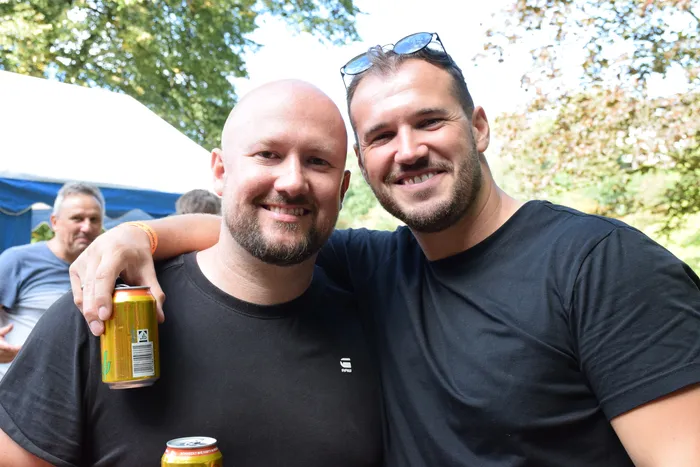 Zwei lächelnde Männer in schwarzen T-Shirts stehen eng beieinander, einer hält eine gelbe Getränkedose.