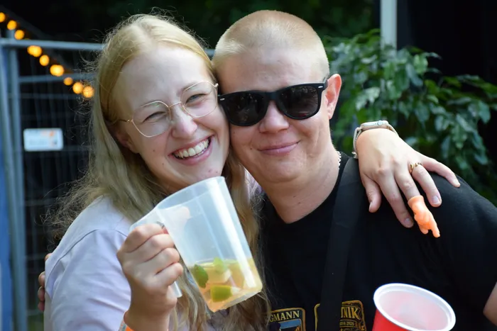 Eine lachende Frau mit Brille und ein Mann mit Sonnenbrille umarmen sich. Die Frau hält einen Krug mit einem Getränk.