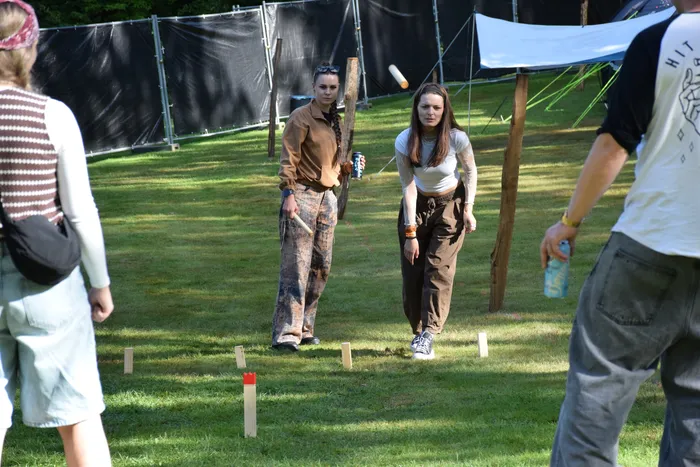 Zwei Frauen spielen ein Holzspiel auf einer Wiese auf einem Campingplatz, während ein Holzstab durch die Luft fliegt.