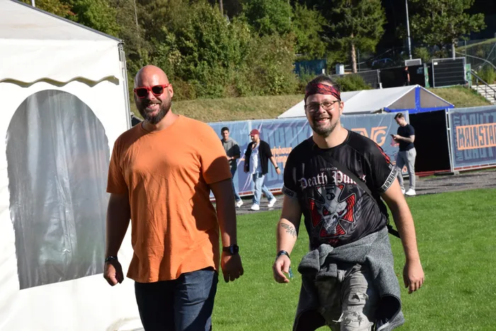 Zwei Männer gehen über eine Wiese auf dem Bautz-Festival. Einer trägt ein orangefarbenes T-Shirt, der andere ein schwarzes T-Shirt und ein rotes Stirnband.