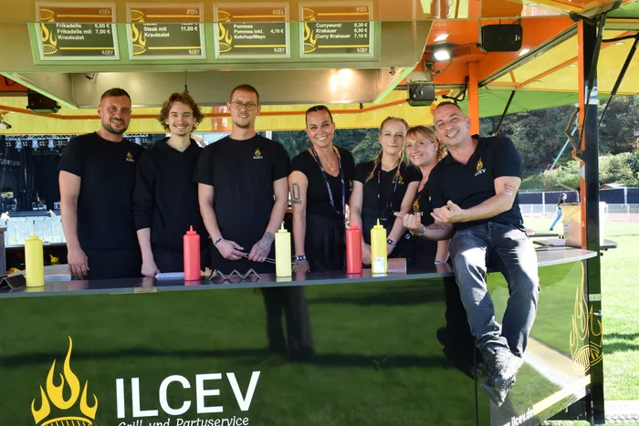 Sieben lächelnde Mitarbeiter des ILCEV Grill- und Partyservice stehen hinter ihrem Verkaufsstand auf dem Bautz-Festival.