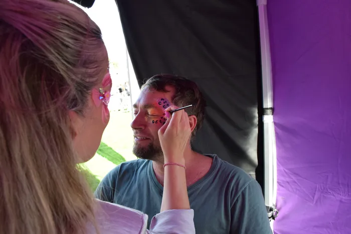 Eine Person trägt mit einem Pinsel bunte Gesichtsbemalung auf das Gesicht eines Mannes auf dem Bautz-Festival auf.