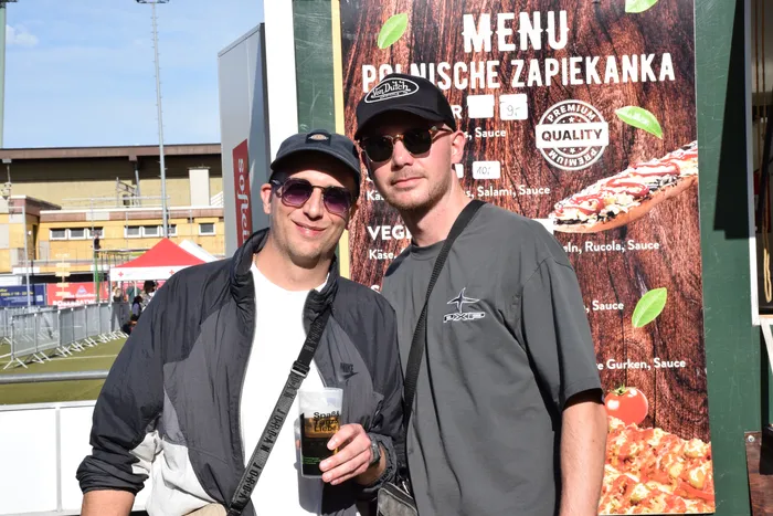 Zwei Männer mit Sonnenbrillen und Kappen posieren auf dem Bautz-Festival vor einem Menüstand für polnische Zapiekanka.