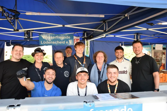 Eine Gruppe lächelnder Menschen posiert hinter einem Tresen unter einem blauen Zelt mit einem Banner der „Wasserfreunde Lüdenscheid e.V.“.