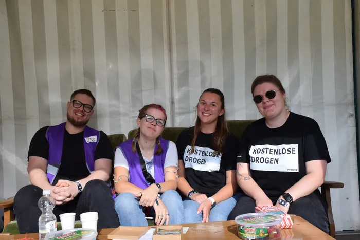 Vier Personen sitzen auf einem Sofa; zwei tragen lila Westen und zwei schwarze T-Shirts mit der Aufschrift „KOSTENLOSE DROGEN *beratung“.