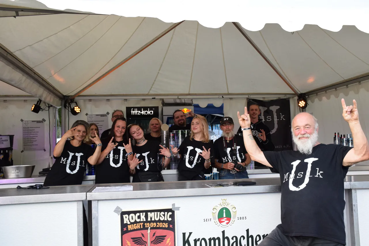 Menschen in schwarzen „Musik“-T-Shirts machen Rock-Handzeichen hinter einem Tresen bei der „Rock Music Night“ unter einem Festzelt.