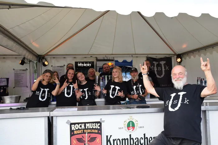 Menschen in schwarzen T-Shirts mit einem 'U'-Logo machen Rock-Handzeichen hinter einer Bar. Ein bärtiger Mann im Vordergrund zeigt beide Hände mit dem Metal-Gruß.