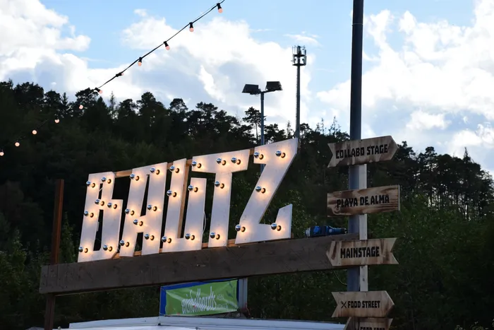 Ein großes, mit Glühbirnen beleuchtetes „BALTZ“-Schild und hölzerne Wegweiser zu verschiedenen Festivalbereichen vor einem Waldhintergrund.