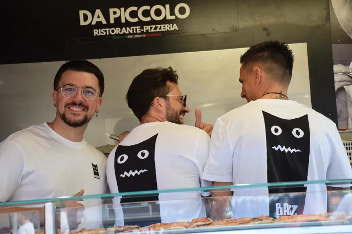 Drei Männer in weißen T-Shirts stehen hinter der Theke des Imbissstands „Da Piccolo“. Zwei zeigen ein schwarzes Katzenmotiv auf dem Rücken.