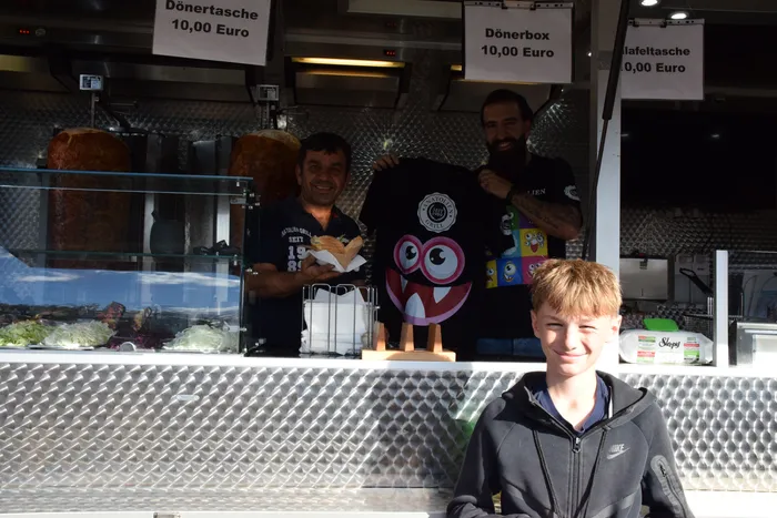 Zwei Männer an einem Imbissstand halten ein Sandwich und ein T-Shirt mit Comic-Motiv hoch, ein Junge lächelt im Vordergrund.
