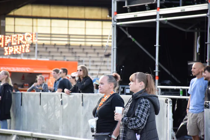 Menschen stehen hinter einer Absperrung auf dem Bautz-Festival vor einer Bühne und einem orangefarbenen Stand mit der Aufschrift „IMPERIAL SPRITZ“.