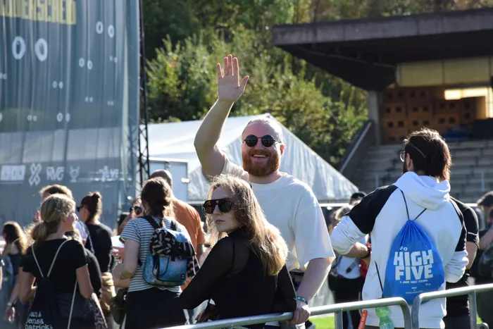 Ein bärtiger Mann mit Sonnenbrille winkt lächelnd in die Menge bei einem Festival.