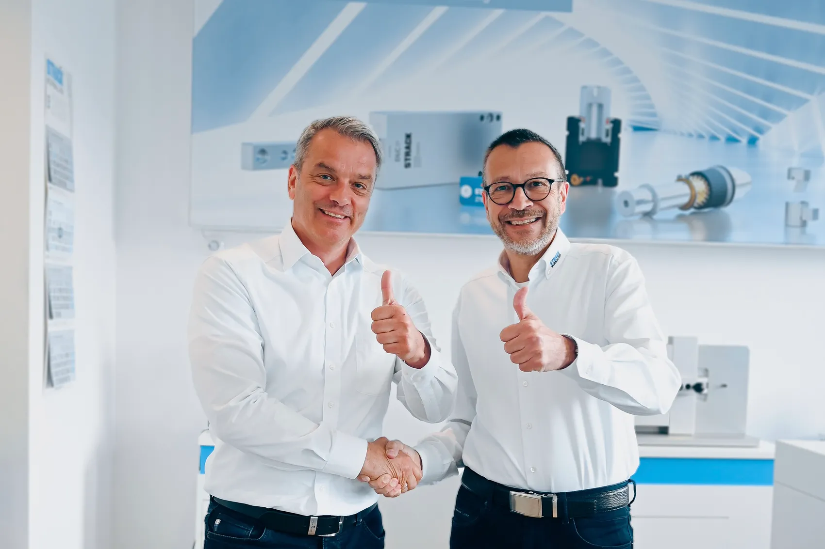 Michael Lang und Andreas Geyer geben sich die Hand und zeigen Daumen hoch, während sie vor technischer Ausrüstung und Plakaten stehen.