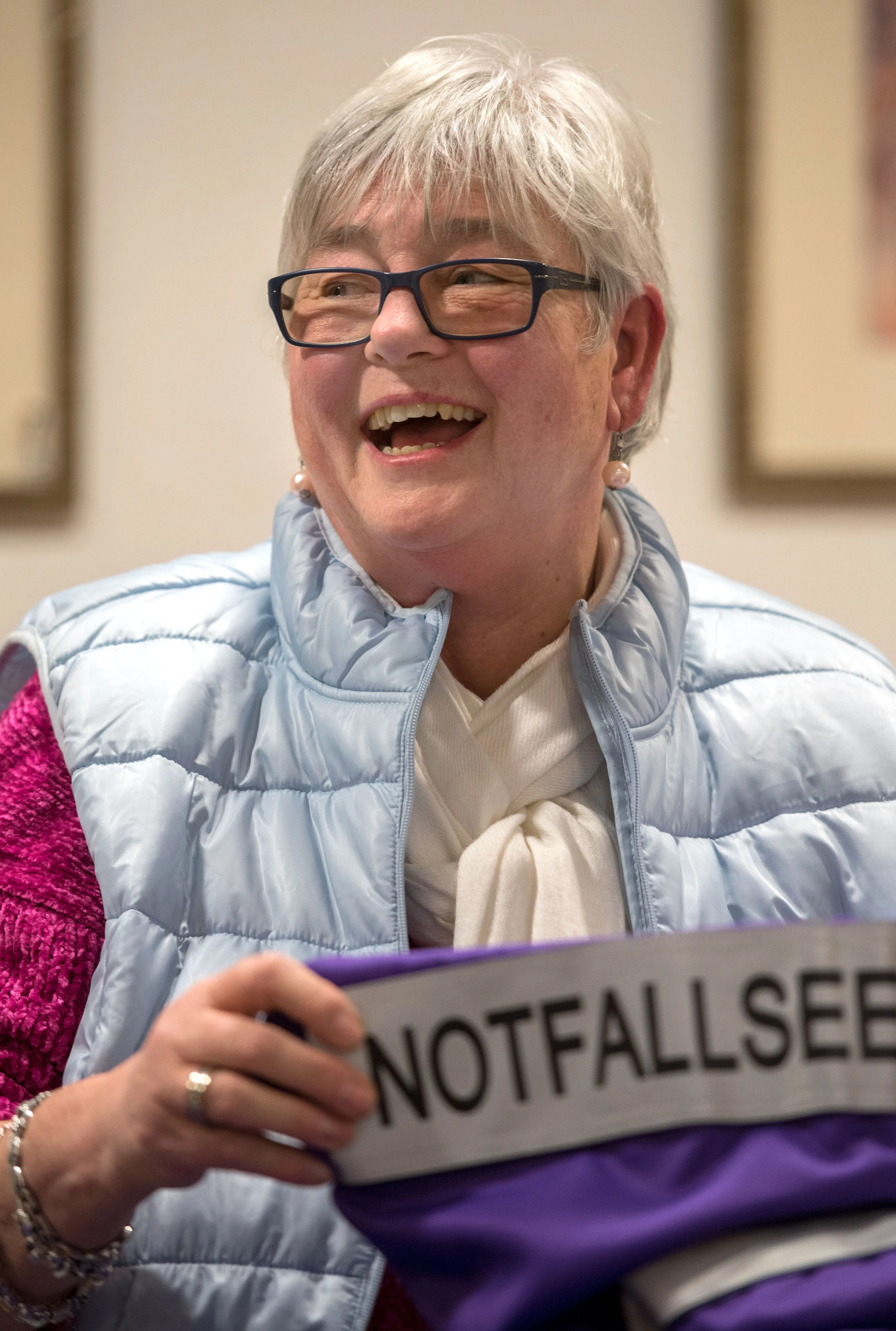 Lachende Frau mit kurzem, grauem Haar und Brille hält ein Schild mit der Aufschrift „NOTFALLSEELSORGE“.