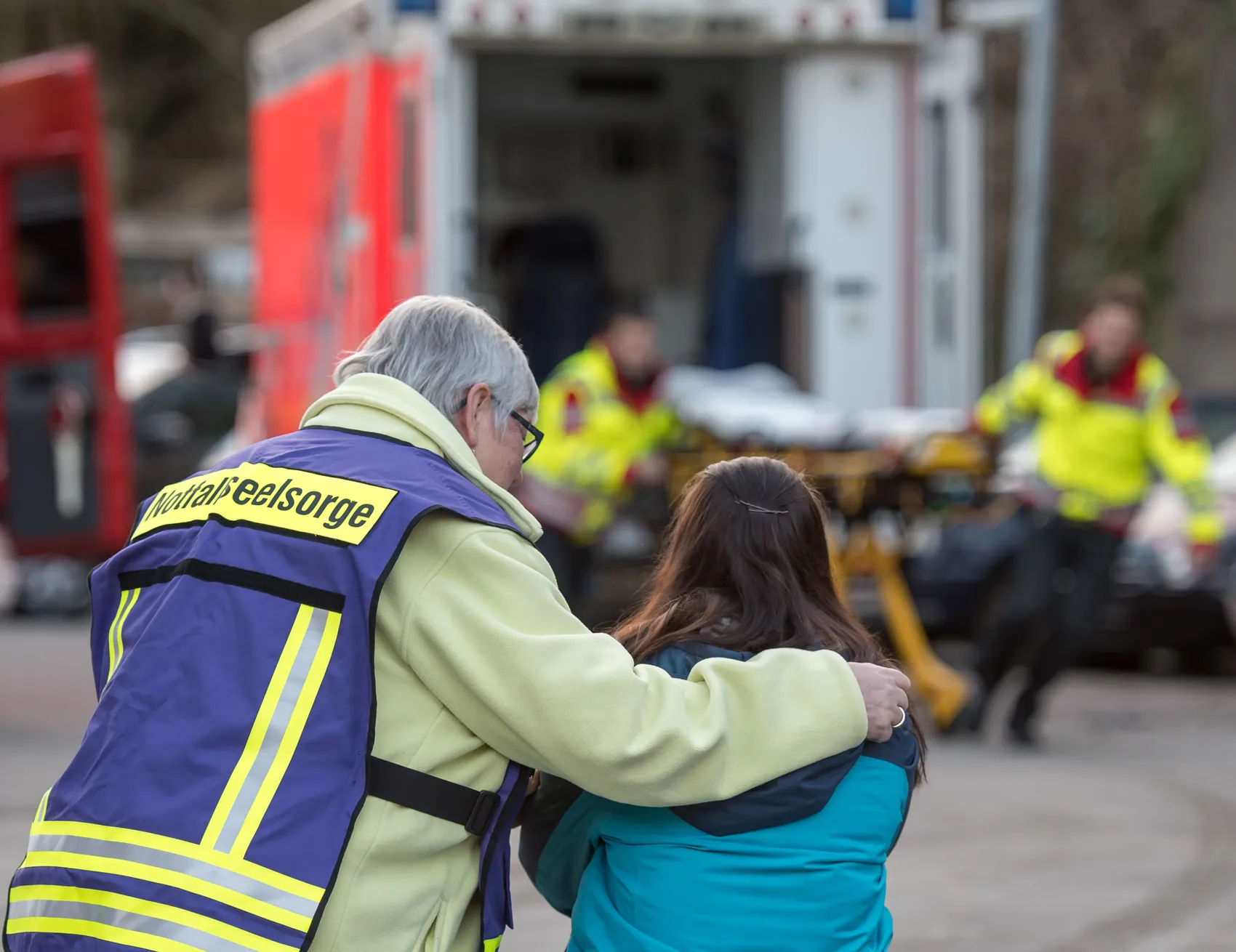 Ein Notfallseelsorger mit gelber Weste umarmt eine junge Frau tröstend, während im Hintergrund ein Rettungswagen und weitere Einsatzkräfte zu sehen sind.