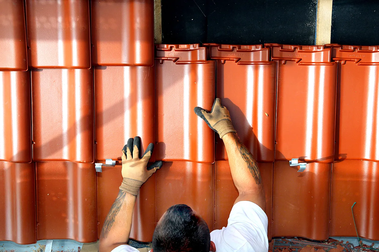 Draufsicht auf einen Dachdecker, der mit behandschuhten Händen orangefarbene Dachziegel verlegt.