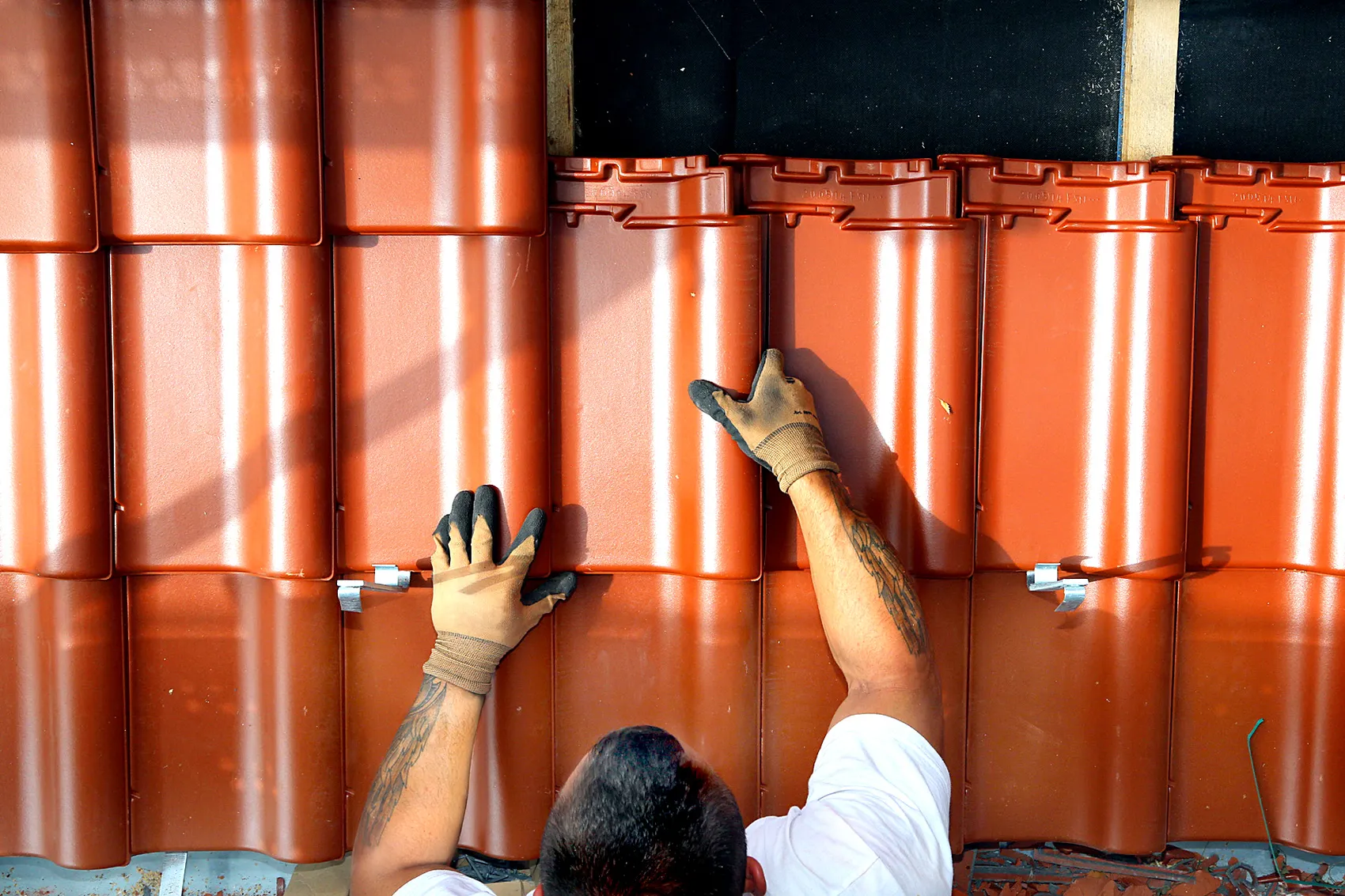 Draufsicht auf einen Dachdecker, der mit behandschuhten Händen orangefarbene Dachziegel verlegt.