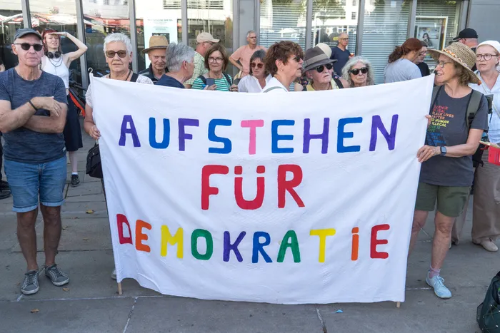 Eine Gruppe von Menschen hält ein weißes Banner mit der bunten Aufschrift „AUFSTEHEN FÜR DEMOKRATIE“.