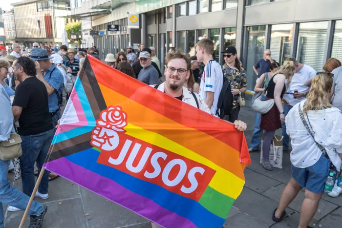 Eine Person hält eine Regenbogenfahne mit dem weißen „JUSOS“-Schriftzug und einem Rosenlogo in einer Menschenmenge.