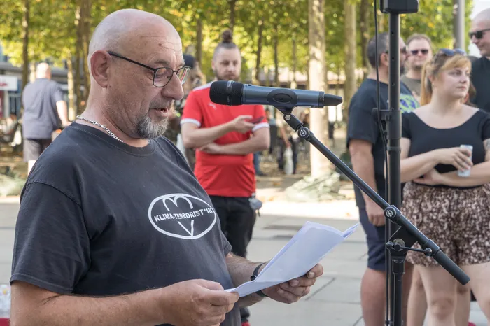 Otto Ersching liest bei einer Kundgebung von einem Papier vor einem Mikrofon. Er trägt ein schwarzes T-Shirt mit der Aufschrift „KLIMA-TERRORIST*IN“.