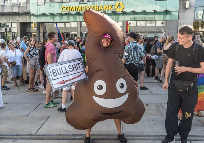 Eine Person im aufblasbaren Haufen-Kostüm hält ein Schild mit der Aufschrift „BULLSHIT“ hoch bei einer Demonstration.
