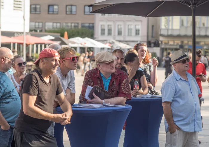Eine Gruppe von Menschen steht an blauen Stehtischen bei einer Kundgebung im Freien.