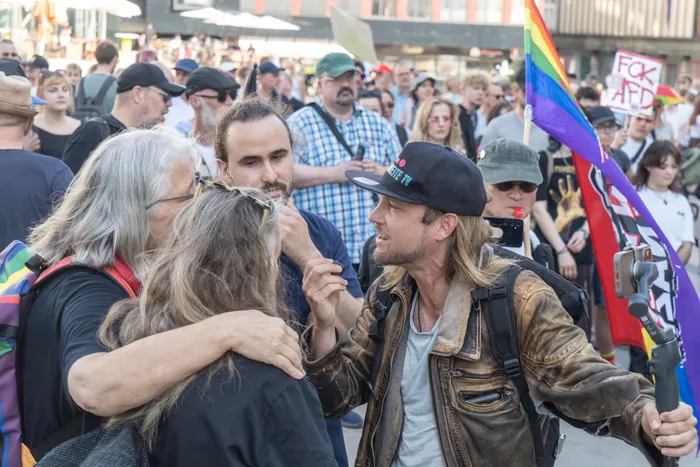 Sebastian Weber von „Weichreite“ TV im Gespräch mit Dr. Gudrun Bekhofer bei einer Demonstration mit Regenbogenfahne und Protestschildern im Hintergrund.