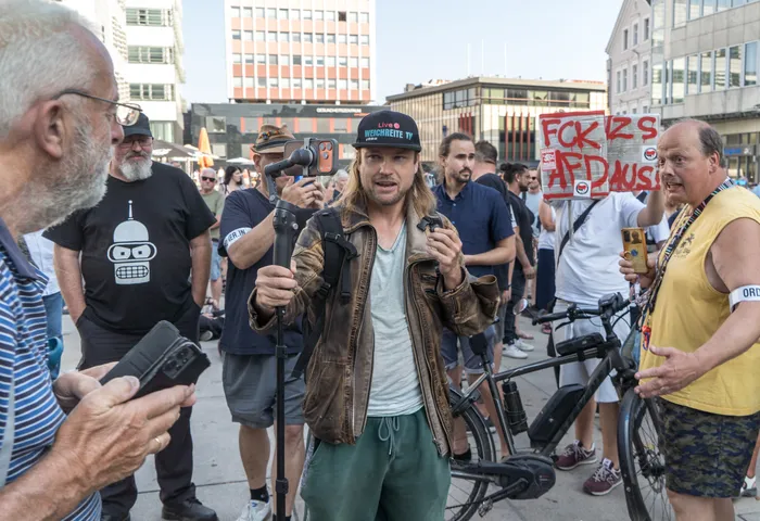 Medienaktivist Sebastian Weber filmt mit einem Smartphone an einem Selfie-Stick; im Hintergrund ist ein Protestschild gegen die AfD zu sehen.