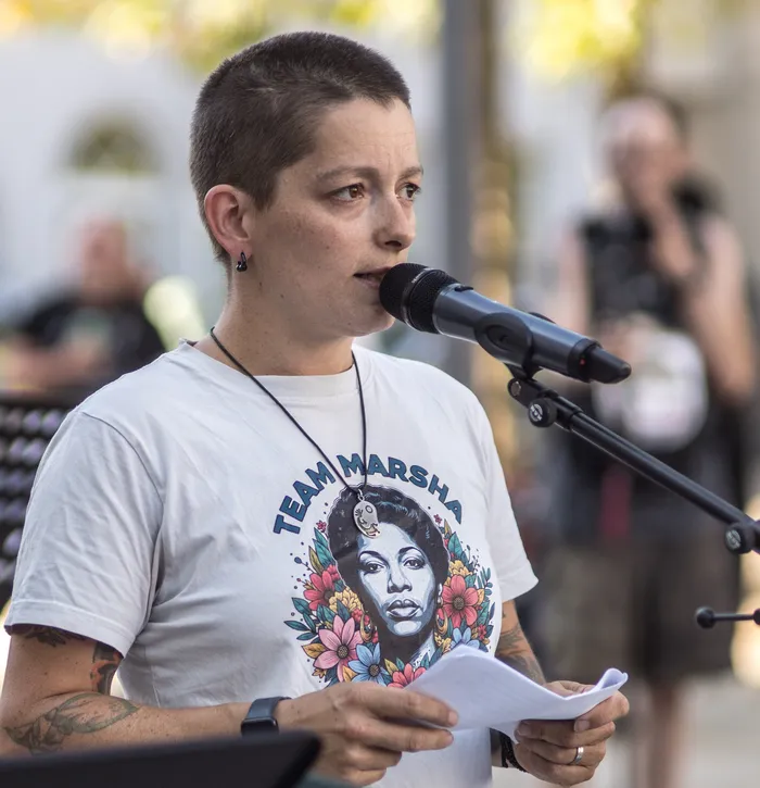 Carina Büdenbender spricht in ein Mikrofon und hält ein Blatt Papier. Sie trägt ein weißes T-Shirt mit einem Porträt und dem Schriftzug „TEAM MARSHA“.